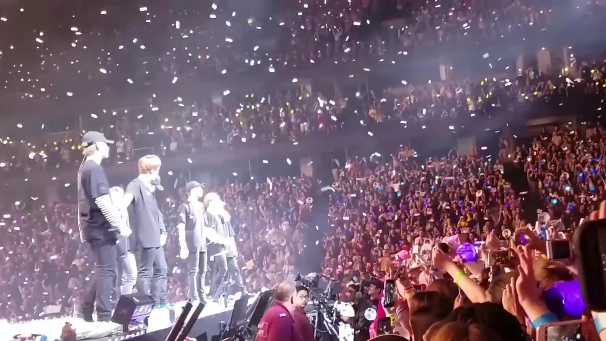 BTS members onstage before a packed arena, with fans holding lights and phones.