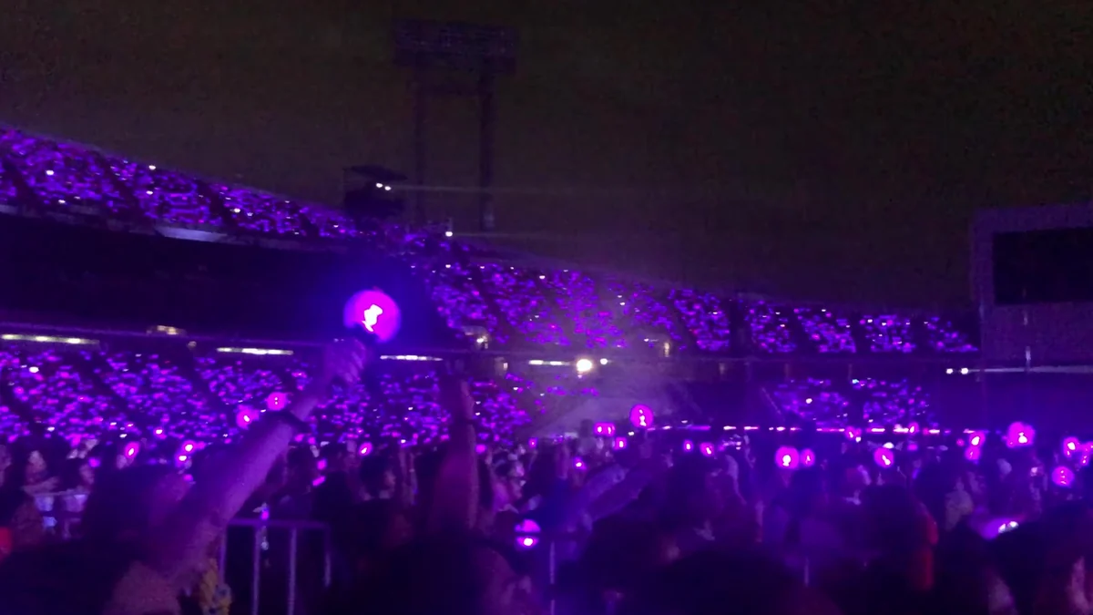 An audience in a dark arena raises purple light sticks during a concert.
