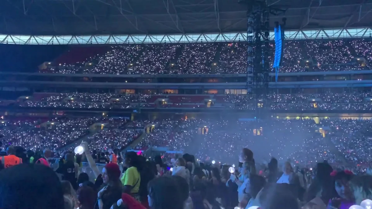 Estádio lotado com milhares de luzes nas arquibancadas e lightsticks no primeiro plano