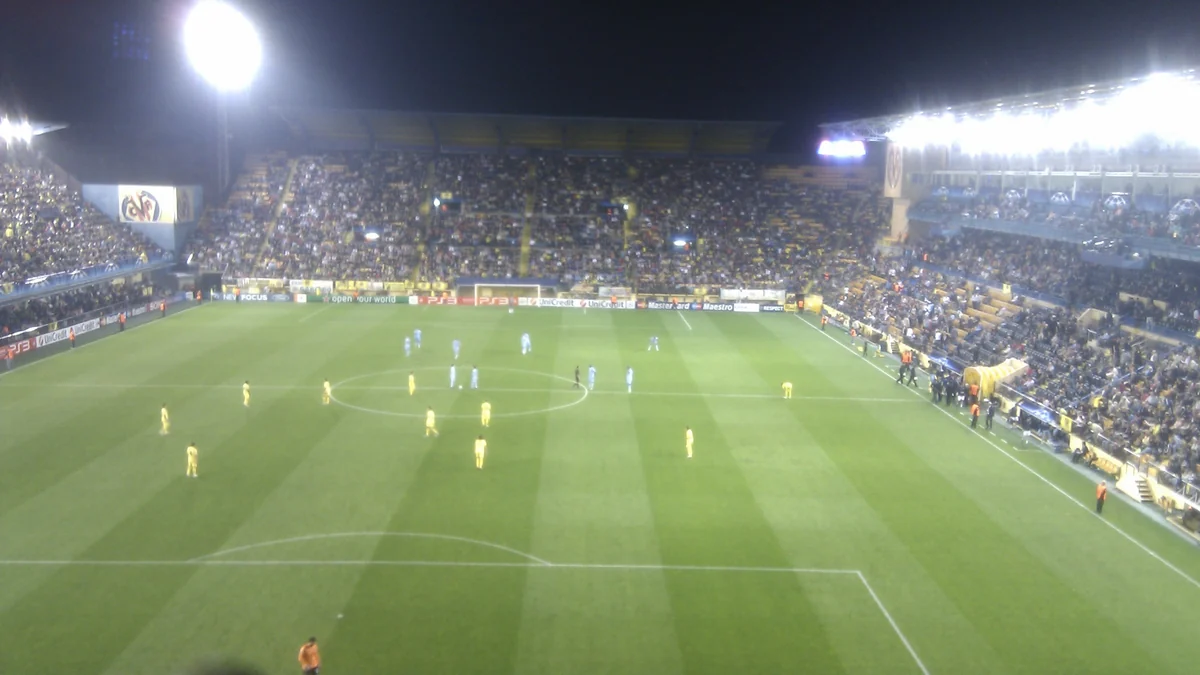 Wide nighttime view of a football pitch with Villarreal players in yellow and opponents in blue