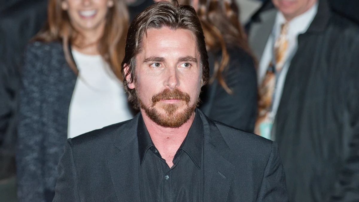 Christian Bale, barbu et vêtu d’un costume sombre, devant des personnes floues