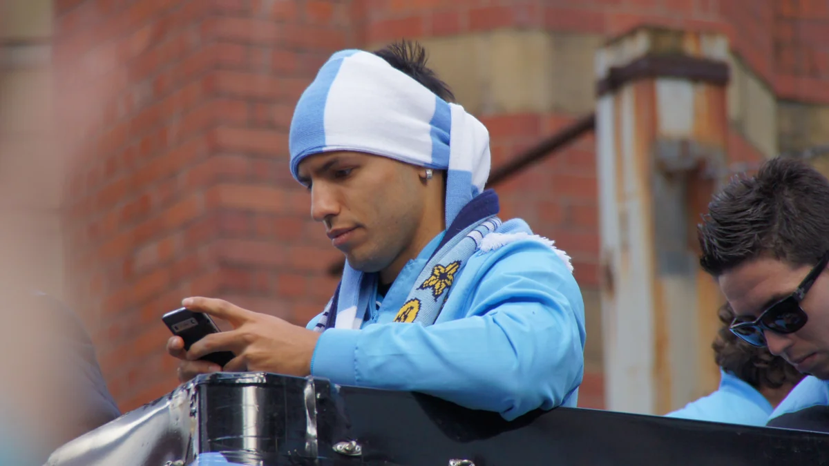 Sergio Agüero wearing a blue-and-white scarf and using a phone on an open-top bus
