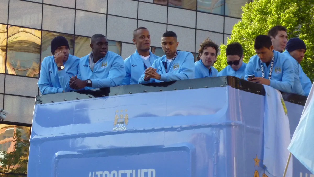 Nine Manchester City players lined up on the upper deck of a blue parade bus