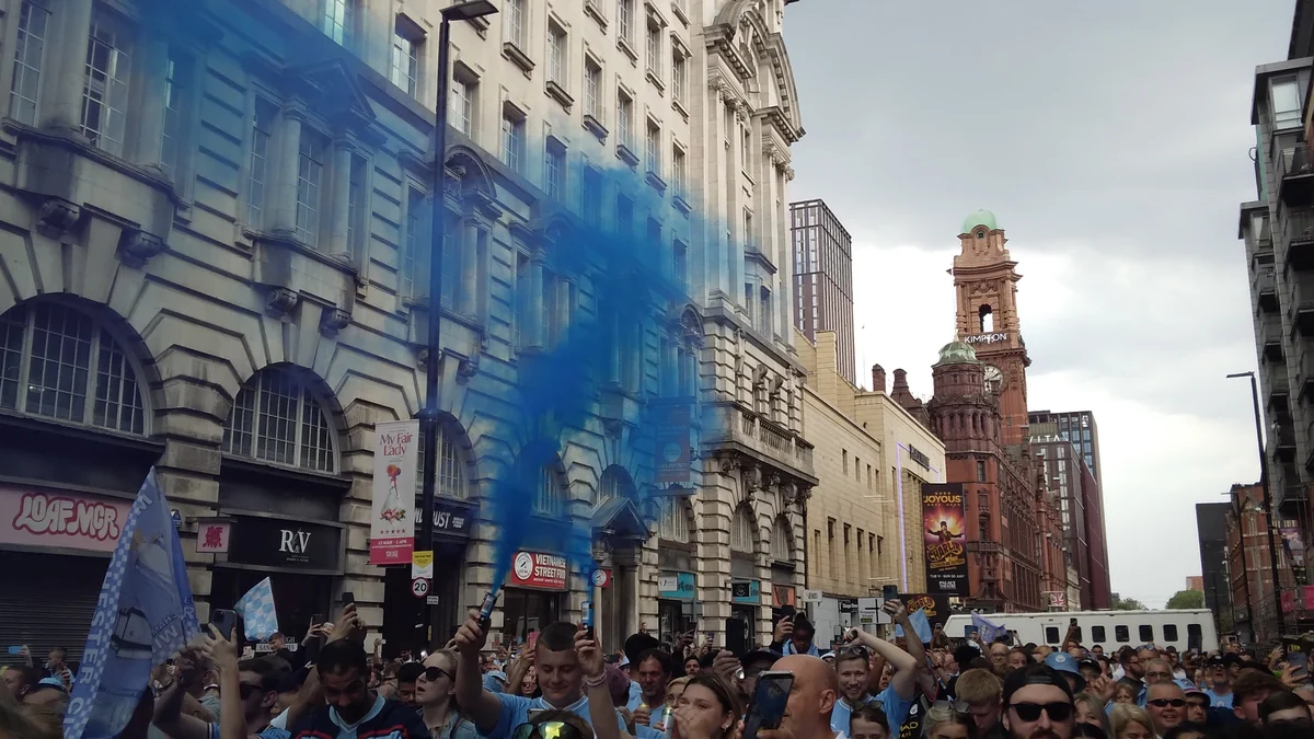 Supporters in light-blue clothing raise phones and a blue smoke flare on a Manchester street