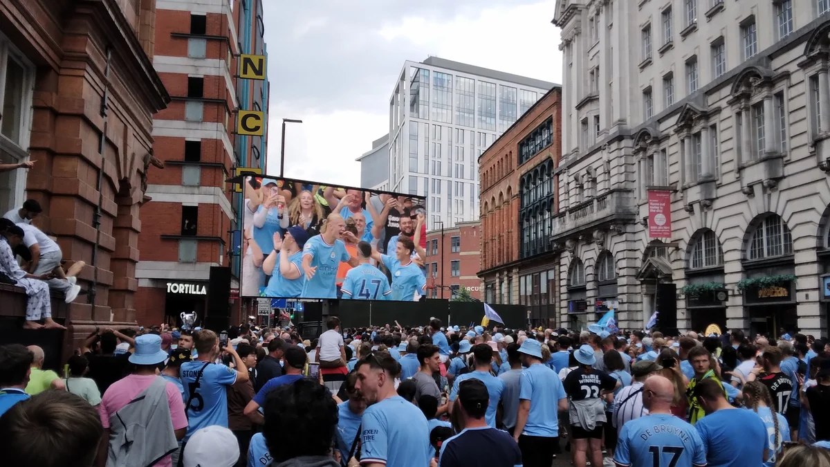 Crowd of Manchester City supporters watching a large screen on a street lined with buildings