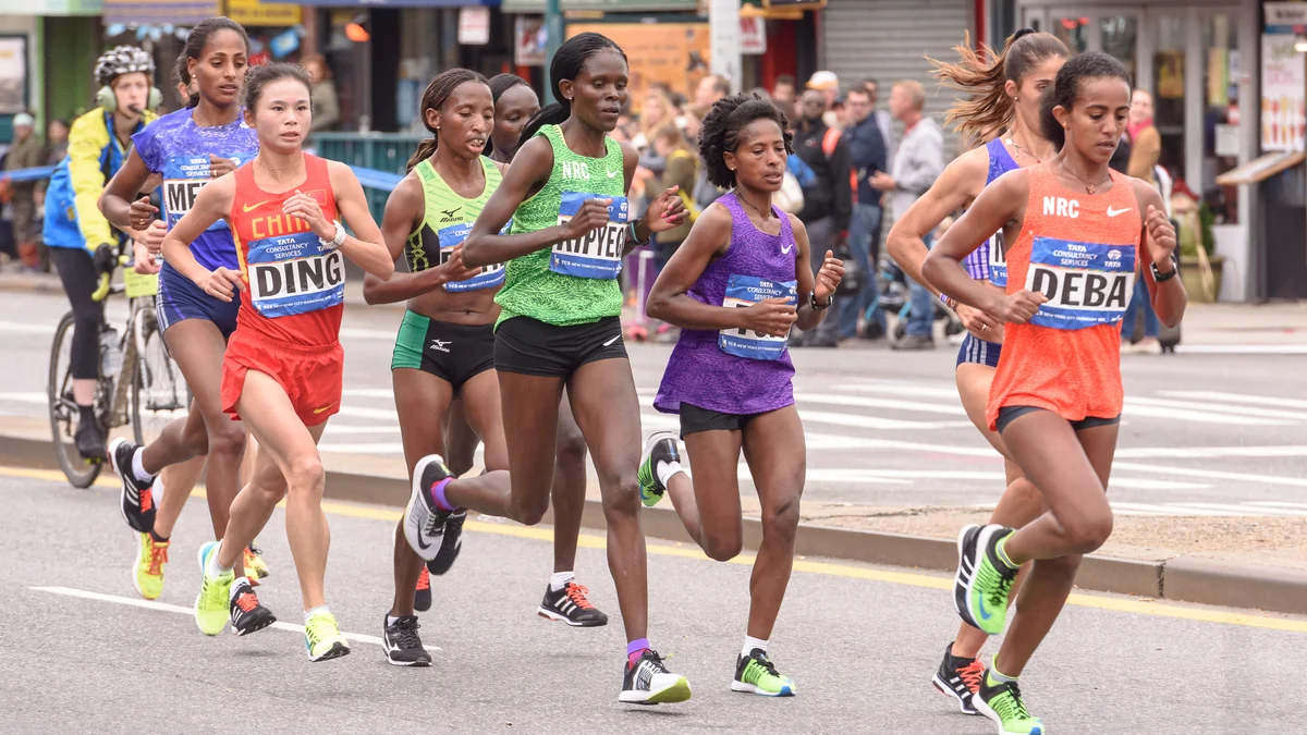 Grupo de corredoras disputa un maratón en una avenida urbana, con público al fondo