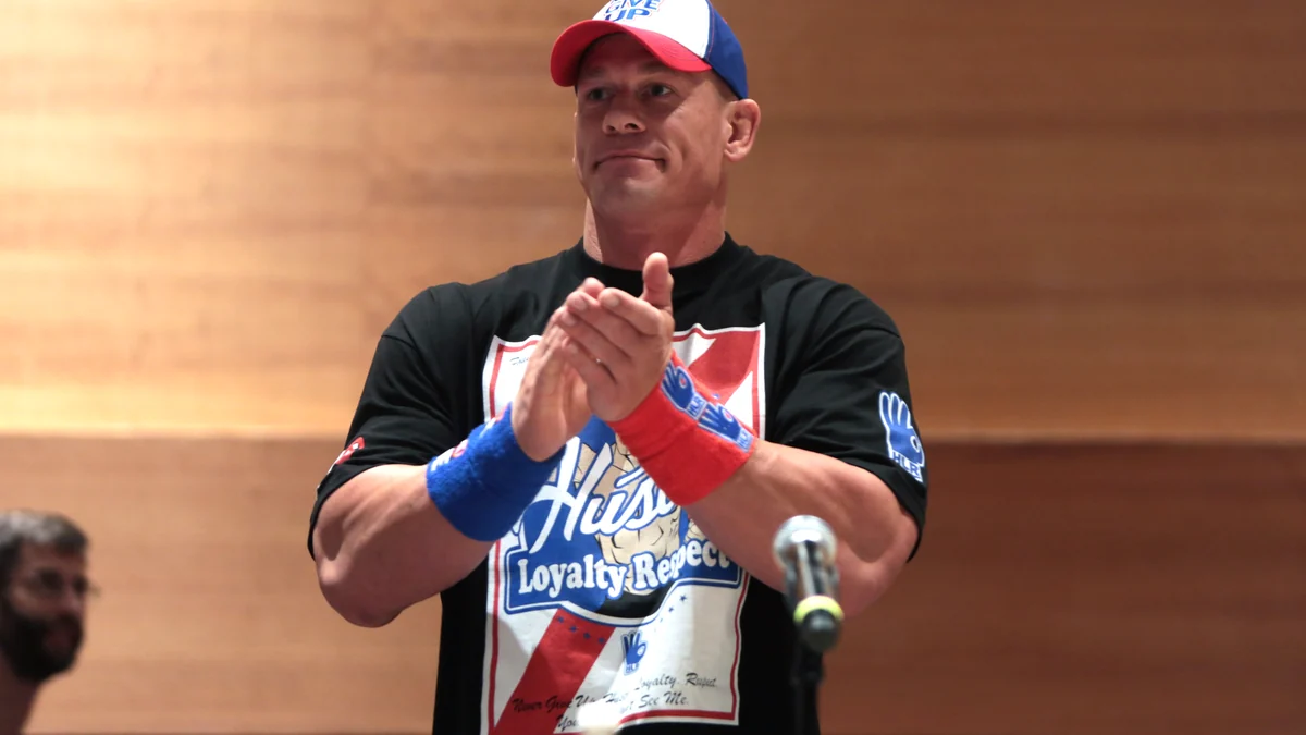 John Cena, con gorra y camiseta negra, aplaude ante un micrófono.
