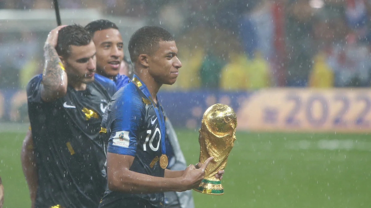 Kylian Mbappé segura o troféu da Copa do Mundo sob a chuva, com medalha no pescoço.