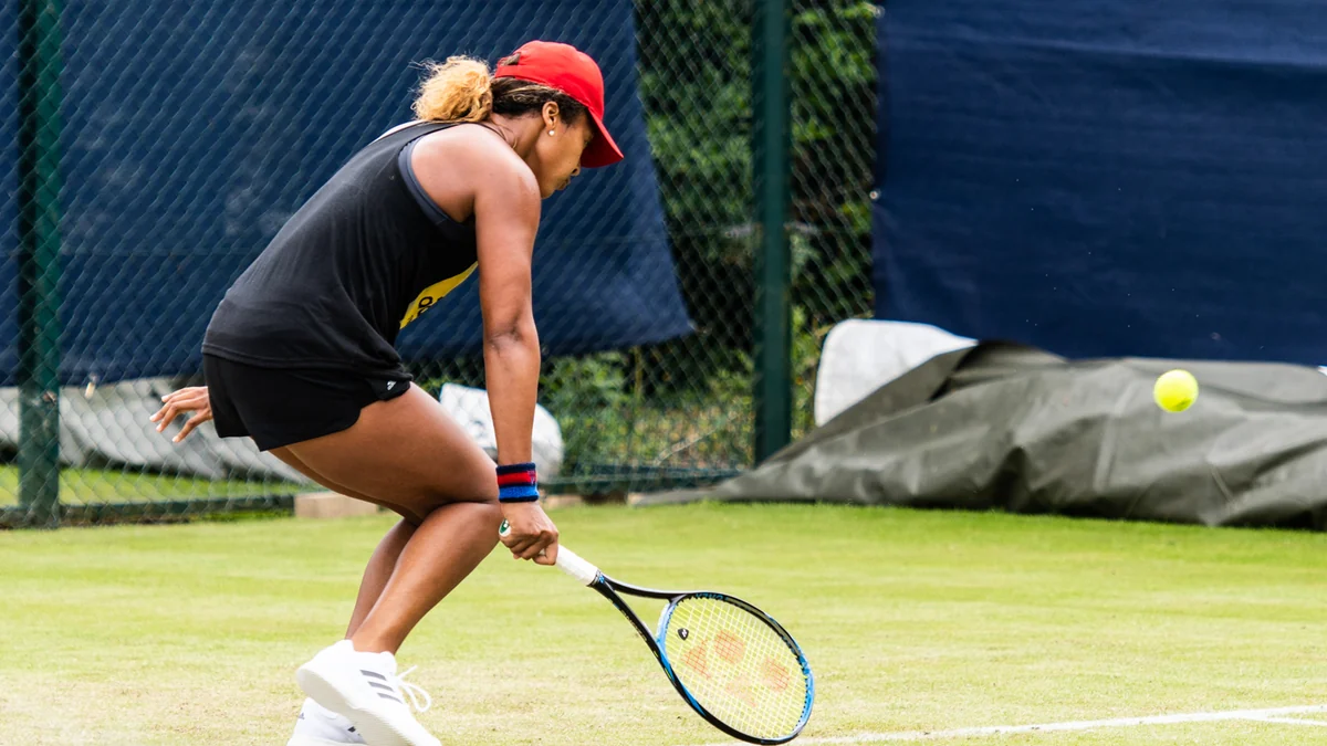 Naomi Osaka, wearing a red cap and black outfit, prepares a low shot on a grass court