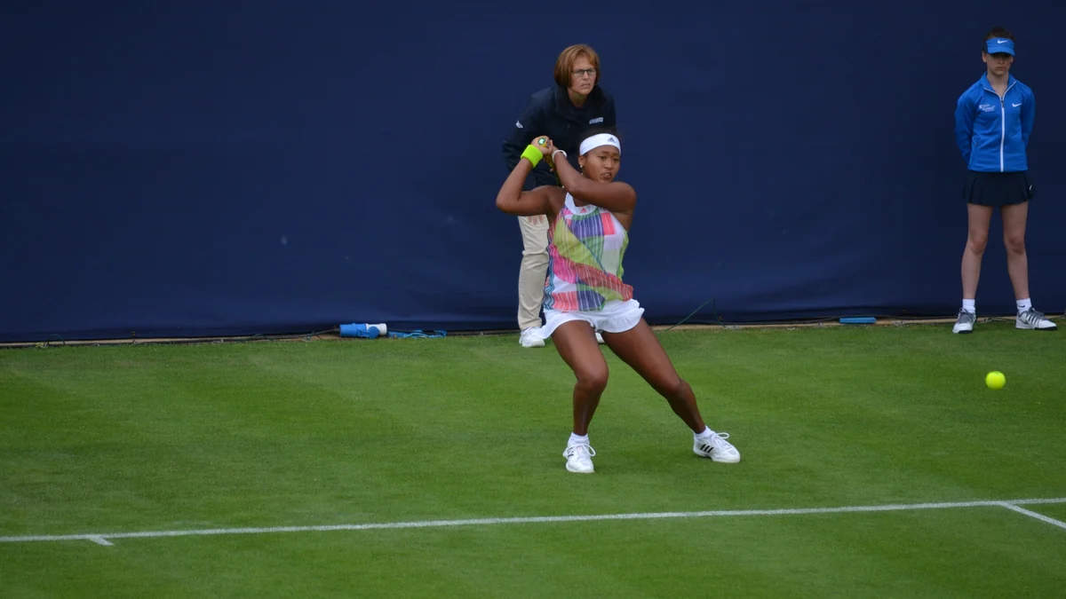 Naomi Osaka hits the ball on a grass court with two people in the background