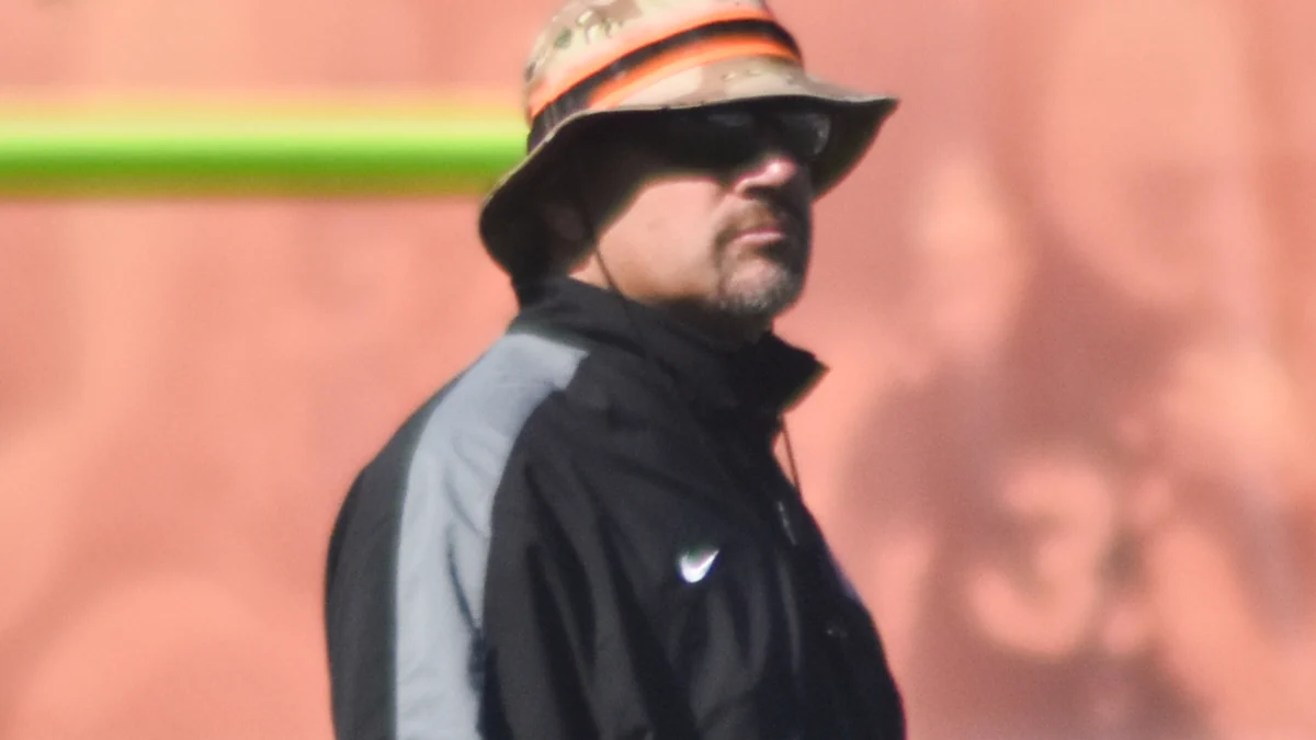 Mike Pettine con sombrero, gafas de sol y chaqueta negra en un campo de entrenamiento