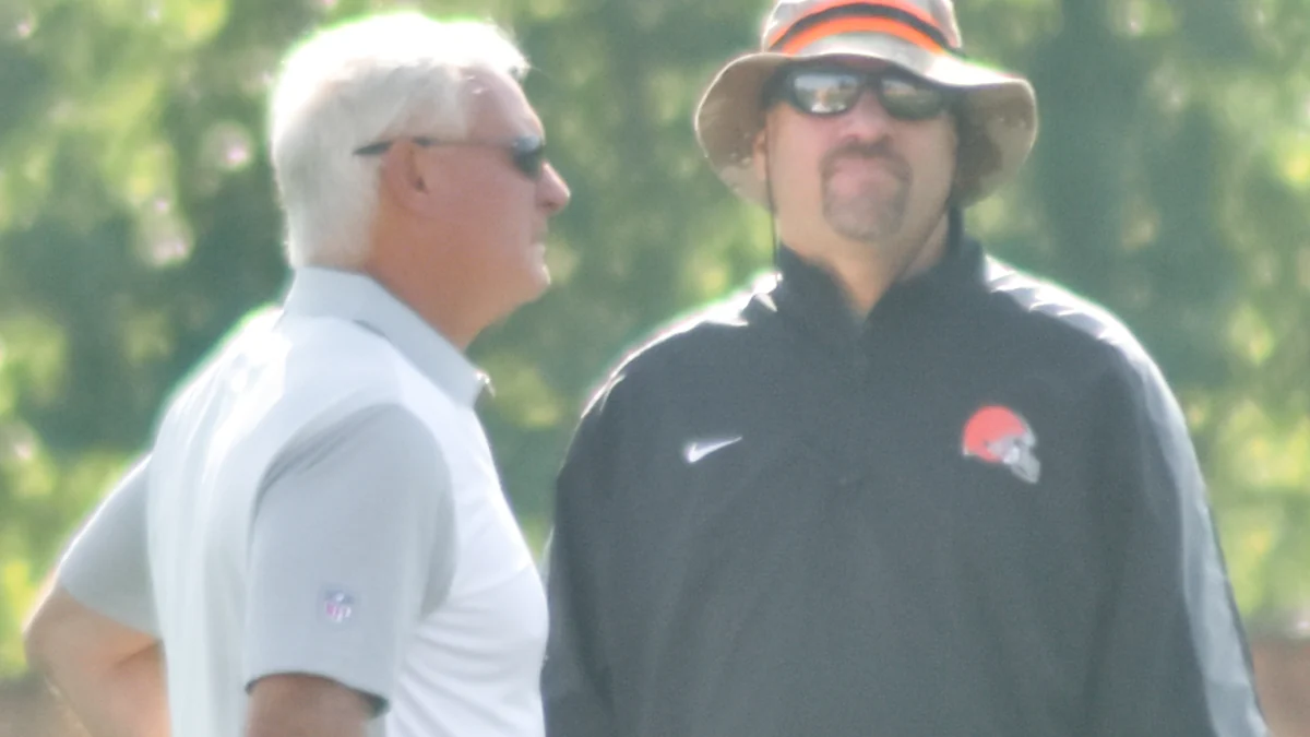 Mike Pettine con sombrero y gafas de sol junto a otro hombre en un campo de entrenamiento