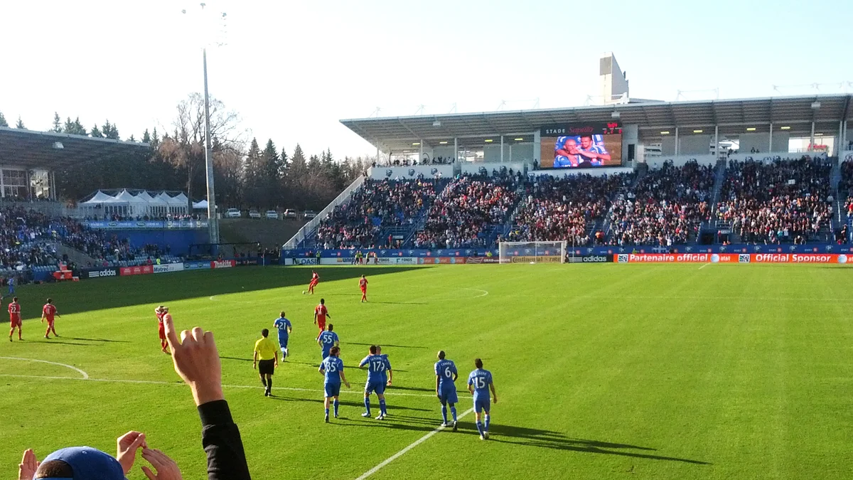 Players in blue and red contest a soccer match in front of occupied stands.