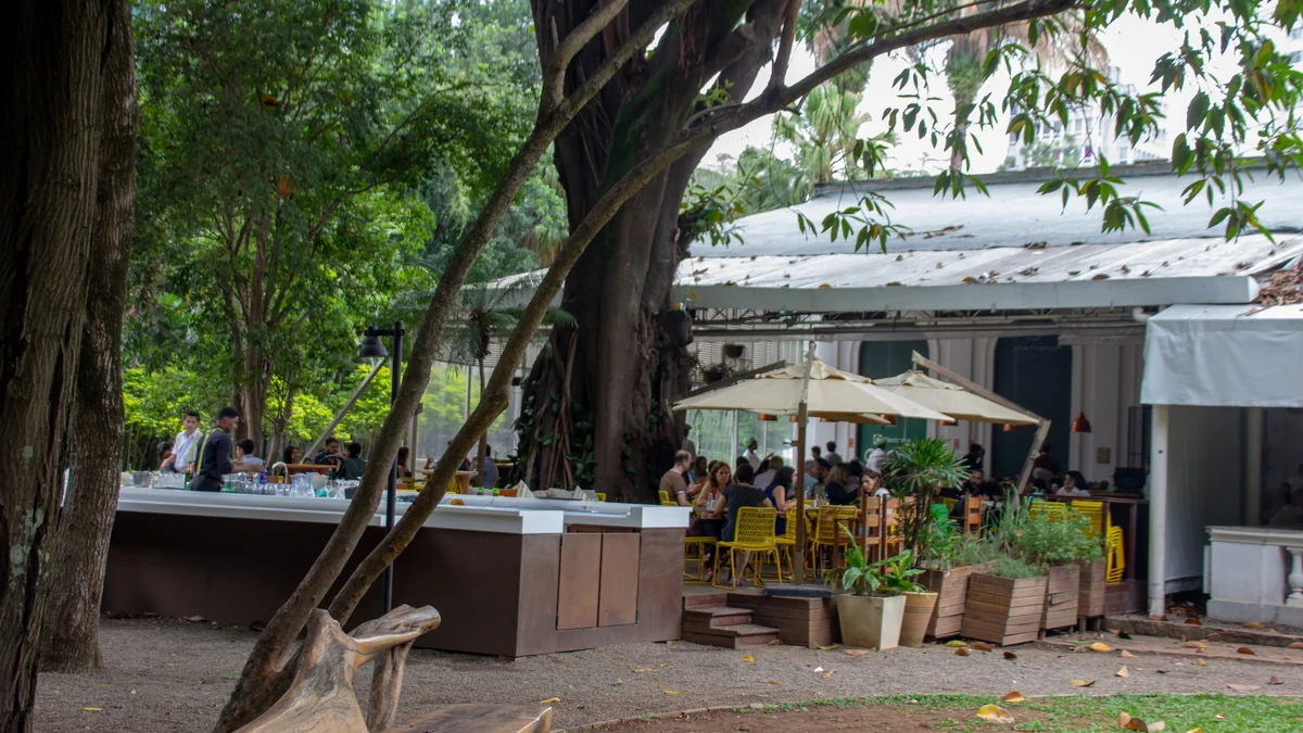 Espace extérieur arboré avec tables, chaises jaunes, parasols et personnes assises