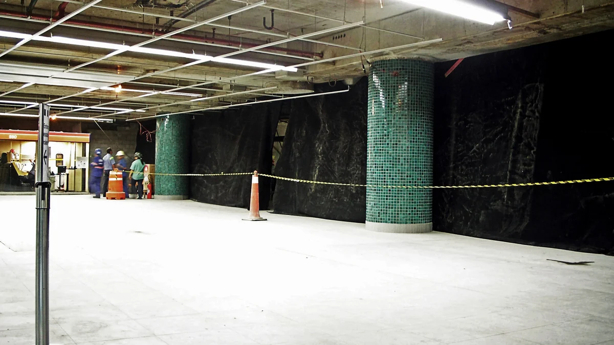 Interior station area under construction, with black barriers, green columns, and caution tape.