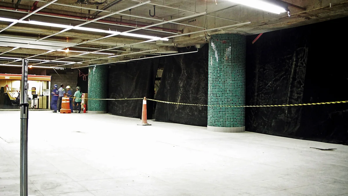 Interior station area under construction, with black barriers, green columns, and caution tape.