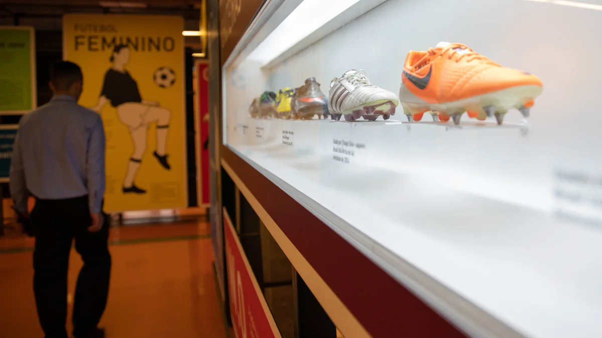 Illuminated display case with football boots and a visitor walking in the museum background.