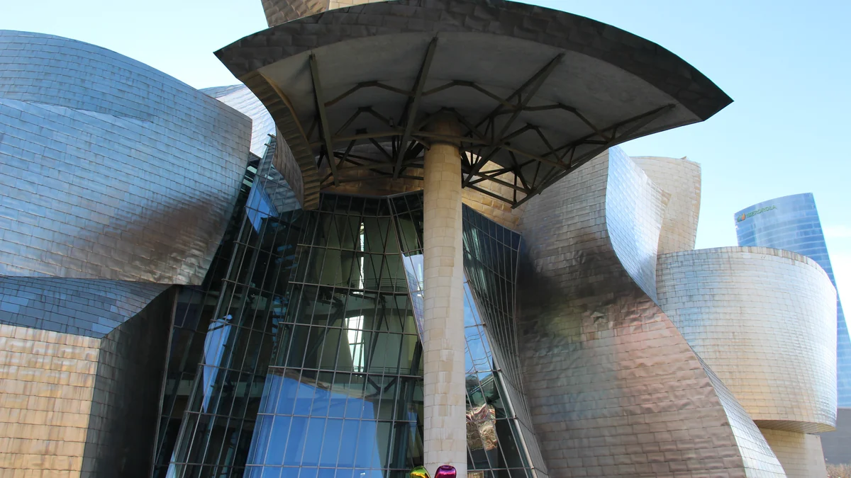 Entrada envidraçada do Museu Guggenheim Bilbao entre volumes metálicos curvos e uma grande cobertura