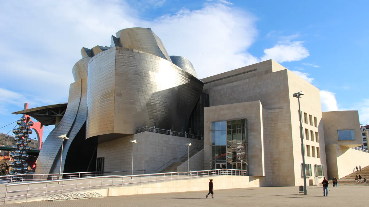 Vista ampla do Museu Guggenheim Bilbao com fachadas metálicas, praça, rampas e algumas pessoas