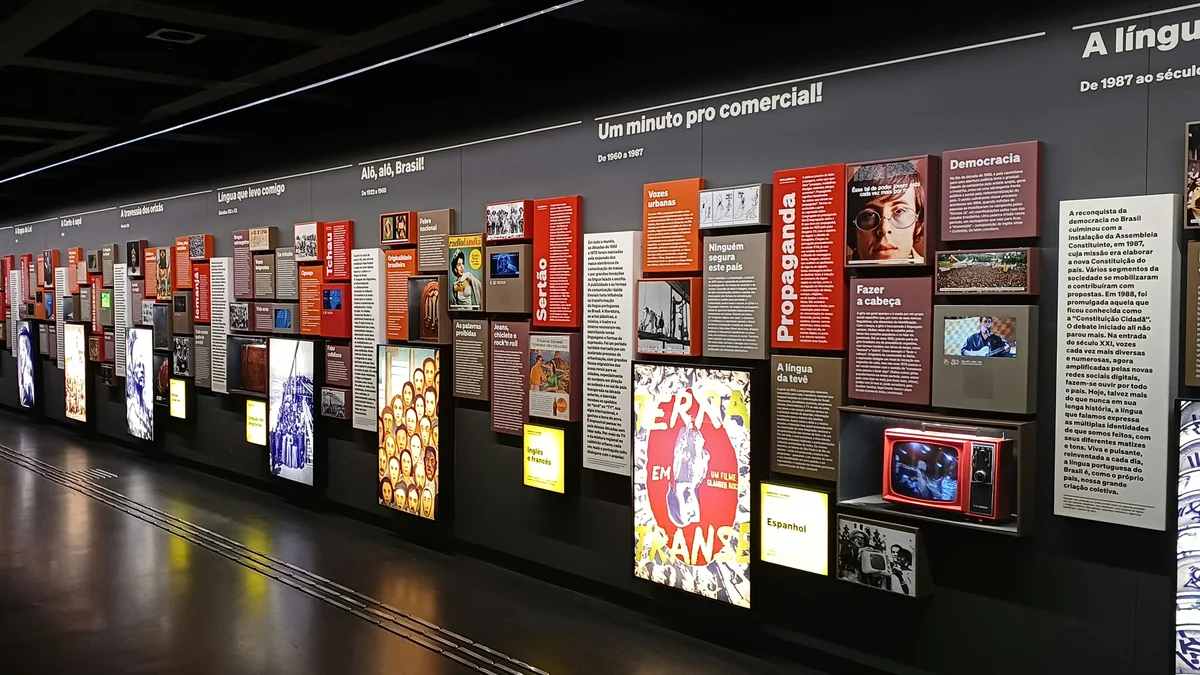 Galeria escura com painéis, fotografias, textos e telas em uma parede expositiva