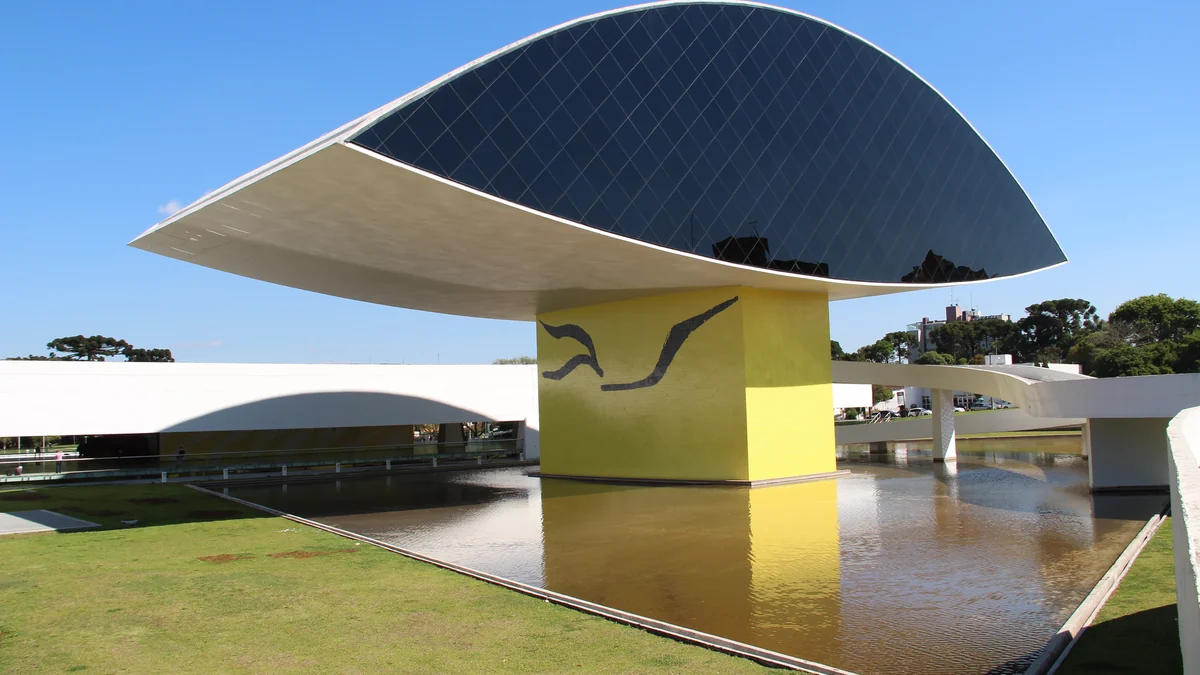 Edifício elevado do Museu Oscar Niemeyer sobre base amarela, diante do espelho d’água