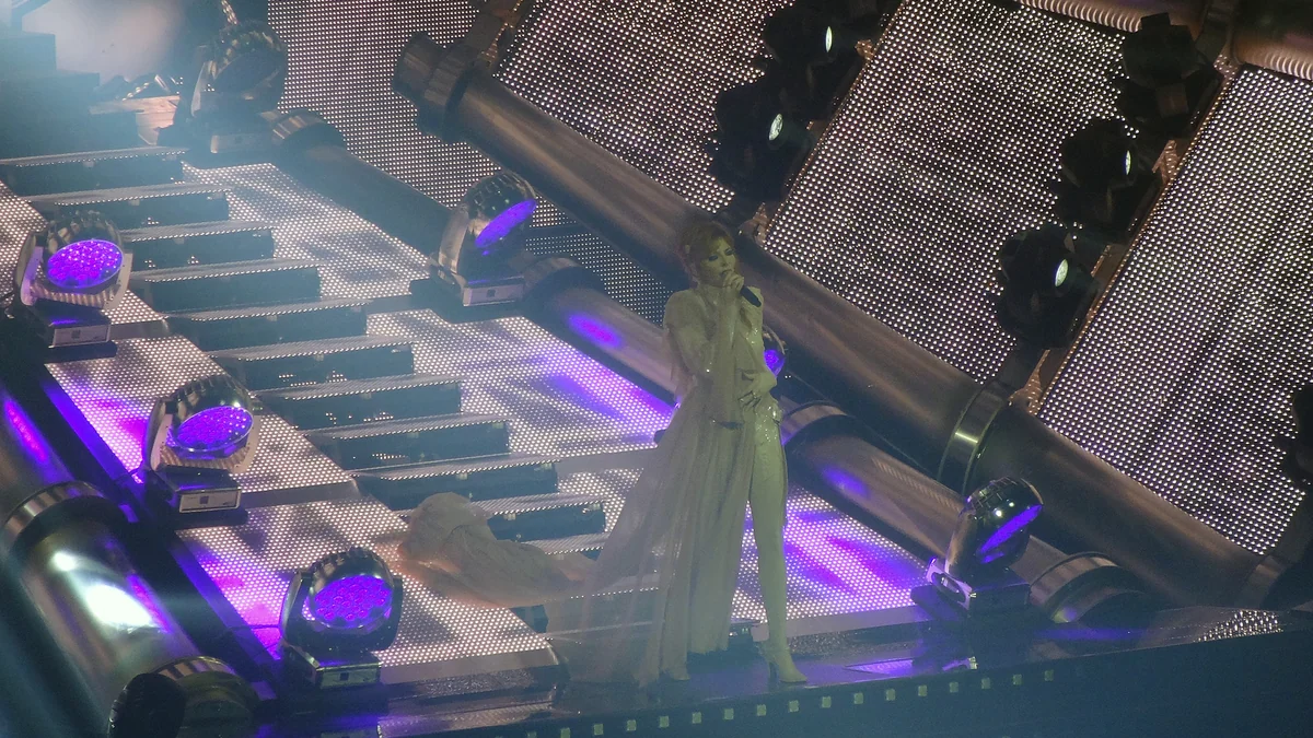 Mylène Farmer in a light-colored costume sings on a runway surrounded by white and violet lights