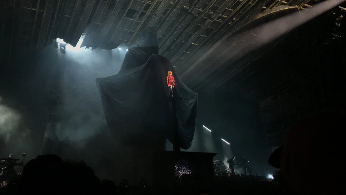 Mylène Farmer in red, suspended before a large dark structure on a smoky stage