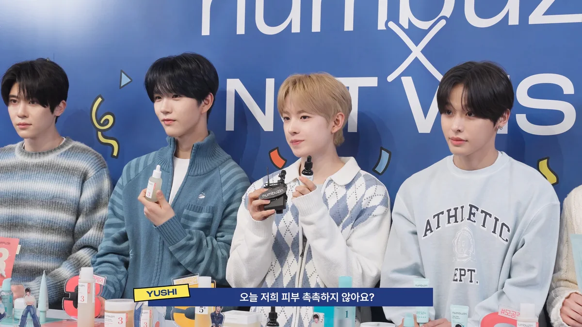 Quatre membres de NCT WISH assis derrière un comptoir avec des cosmétiques, sur fond bleu.