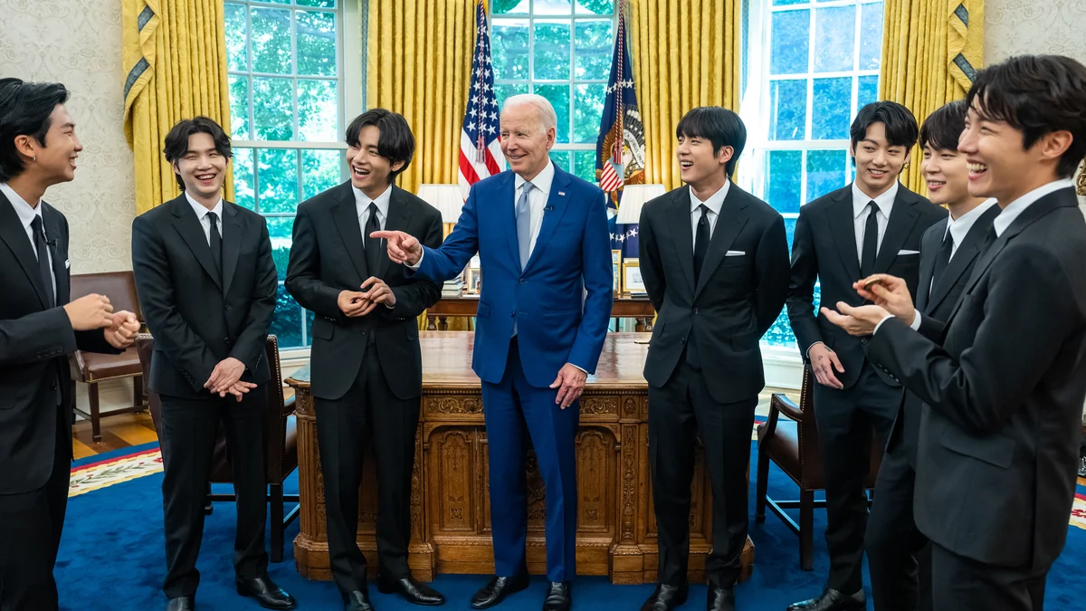 Los siete integrantes de BTS, con trajes negros, conversan y sonríen alrededor de Joe Biden, vestido con un traje azul.