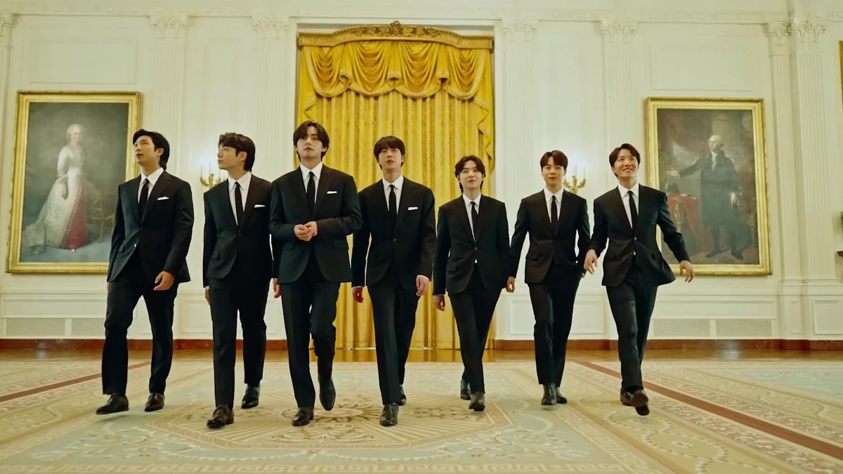 Sete integrantes do BTS caminham em uma sala ampla, usando ternos pretos, diante de cortinas douradas e dois retratos.