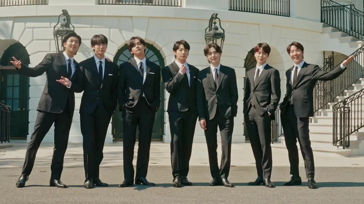 Sete integrantes do BTS posam de terno preto ao ar livre diante de um edifício branco com escadas laterais.