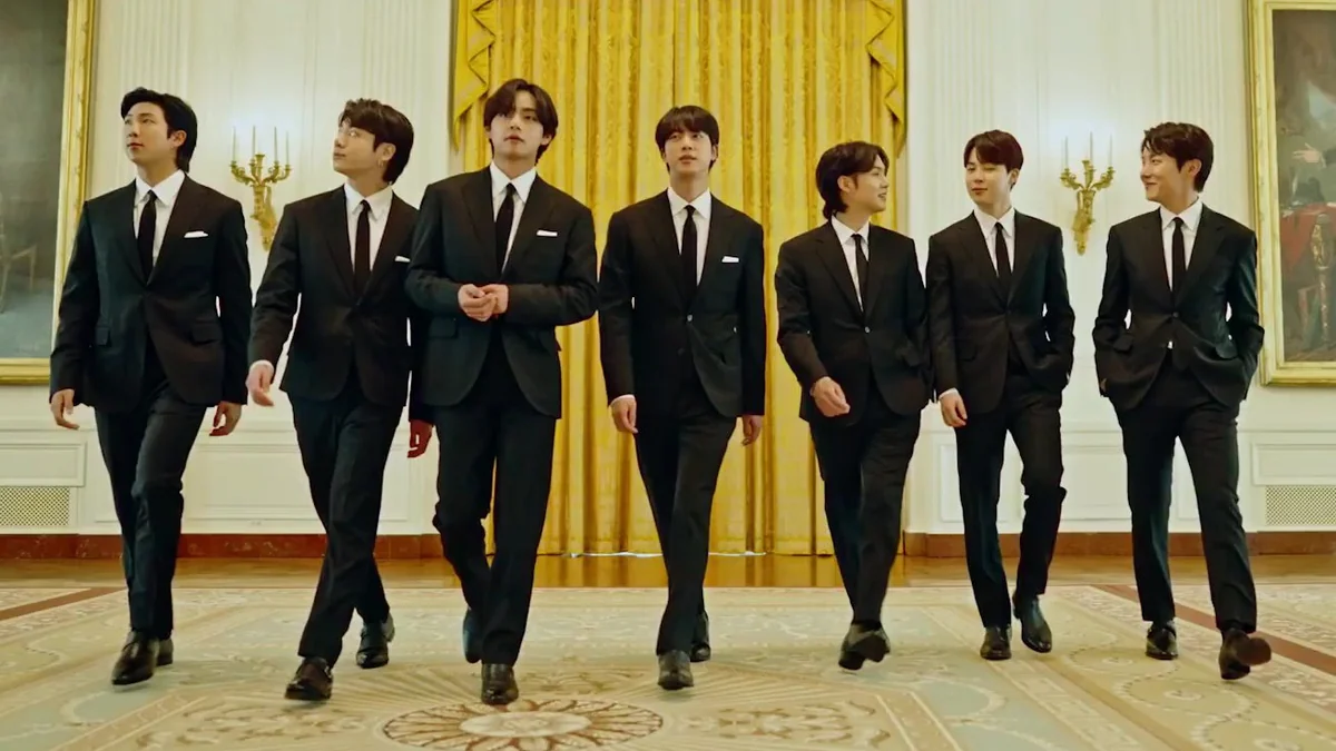 Sete integrantes do BTS caminham de terno preto diante de cortinas douradas em uma sala formal.