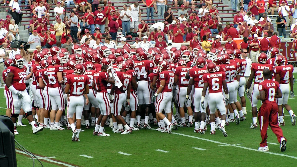 Oklahoma Sooners players gather in a huddle on the field with fans in the stands.