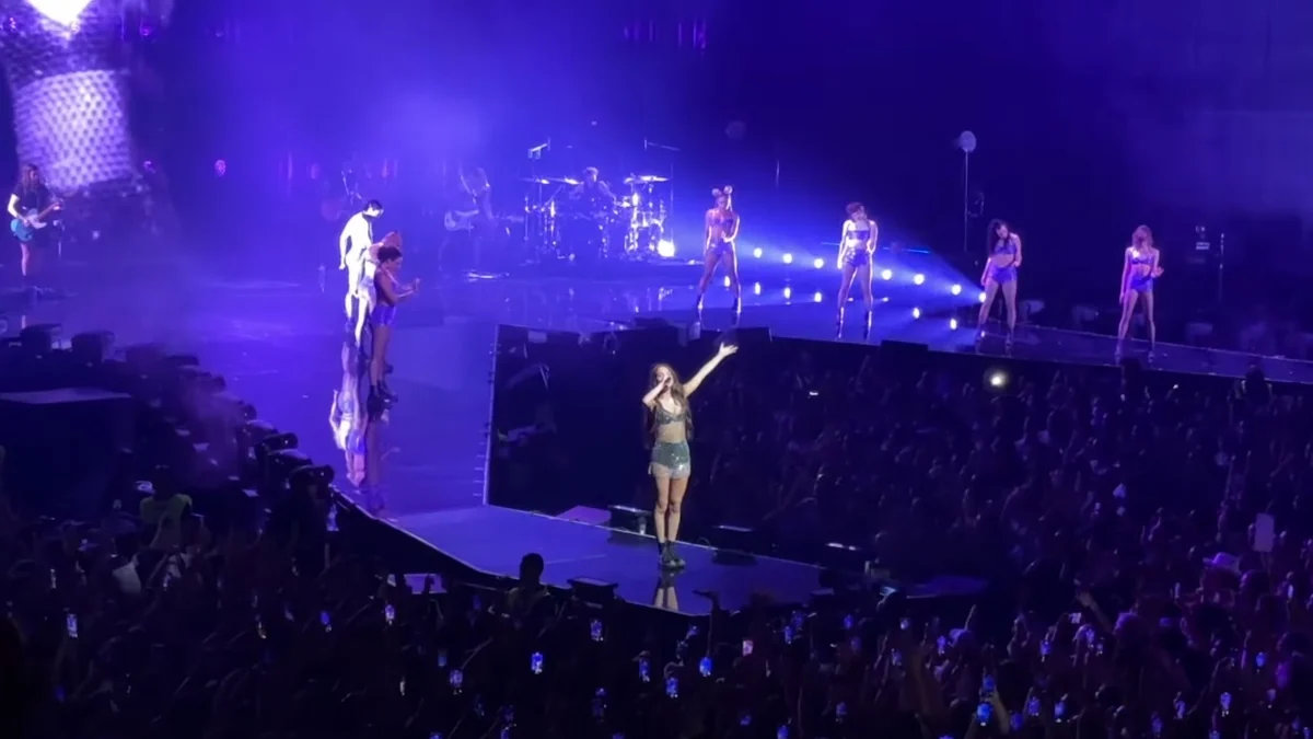 Olivia Rodrigo canta na passarela de um palco com dançarinos e banda ao fundo, diante de uma plateia