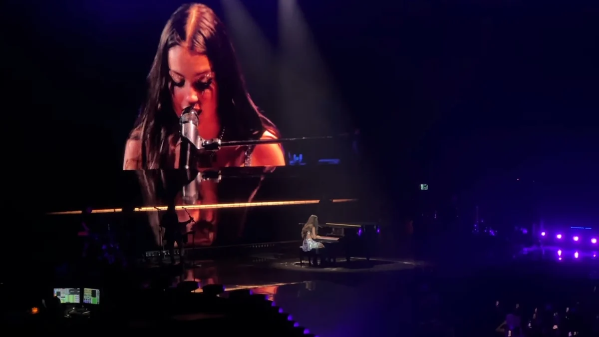 Olivia Rodrigo canta ao piano enquanto seu rosto aparece ampliado no telão do palco