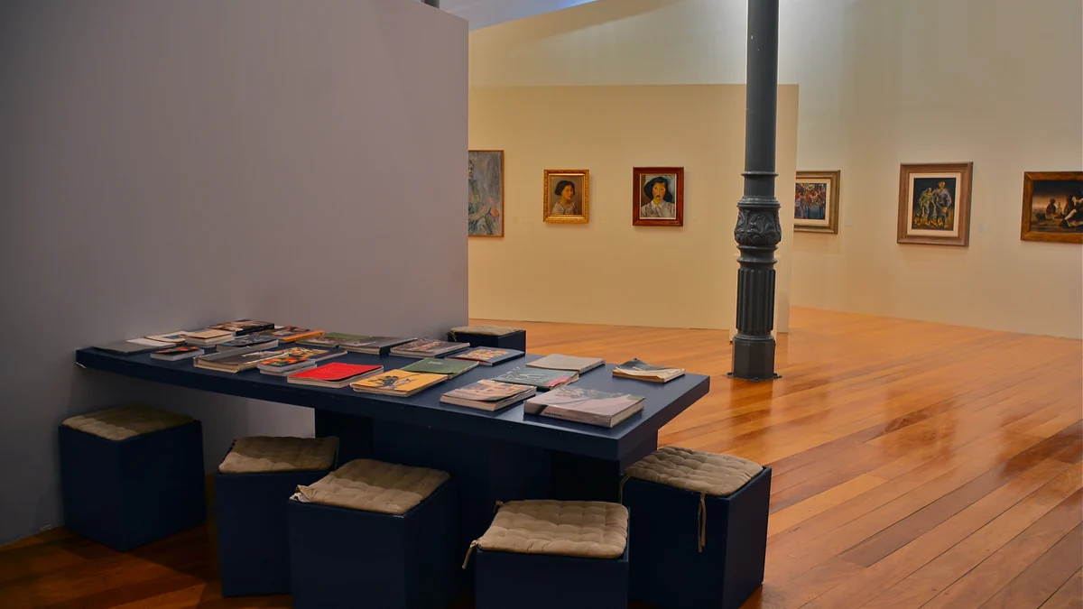 Gallery interior with wooden floor, paintings on the walls and a table covered with publications