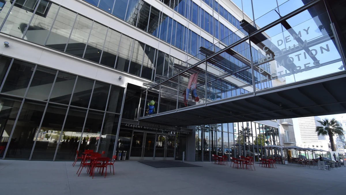 Glass facade of the Academy Museum of Motion Pictures, with an elevated walkway and red tables outside.