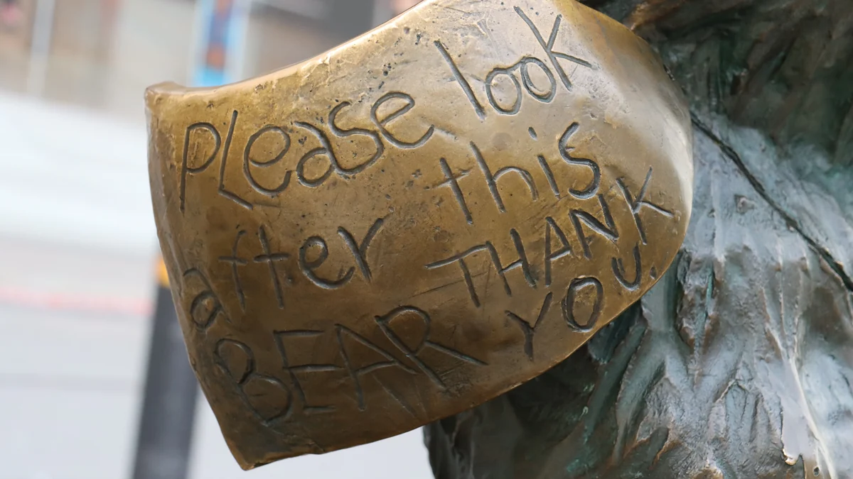 Gros plan sur l’étiquette en bronze de la statue portant la phrase anglaise « Please look after this bear. Thank you »