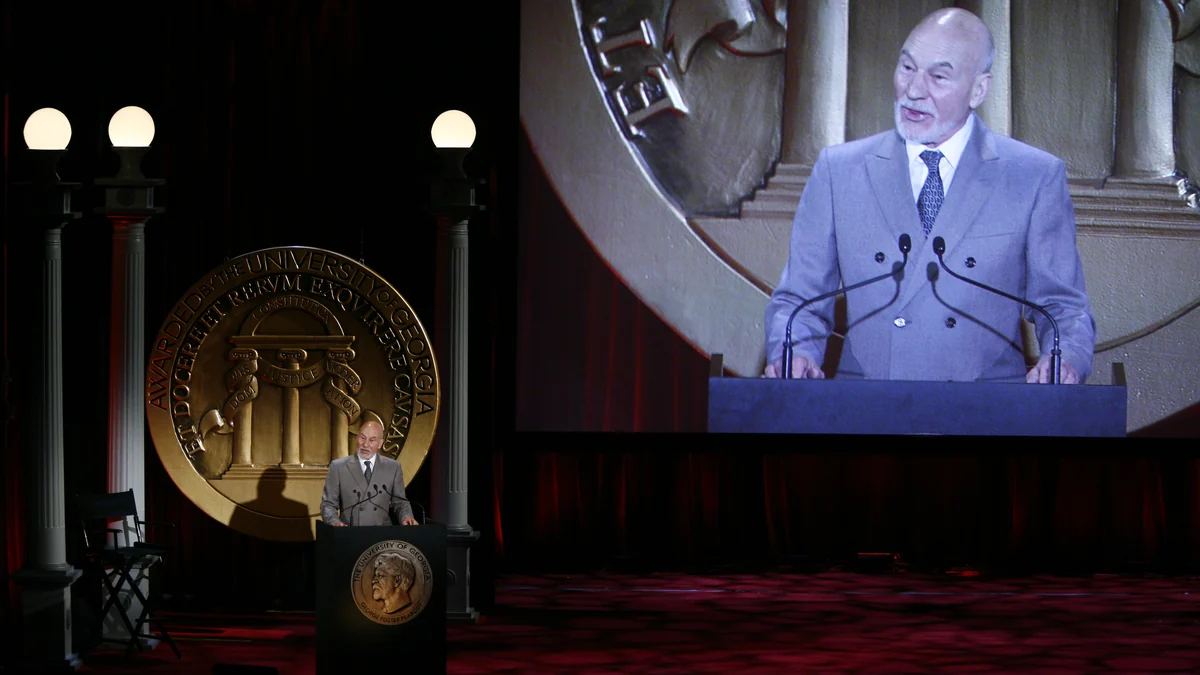 Patrick Stewart parle à un pupitre tandis que son image apparaît sur un grand écran derrière lui