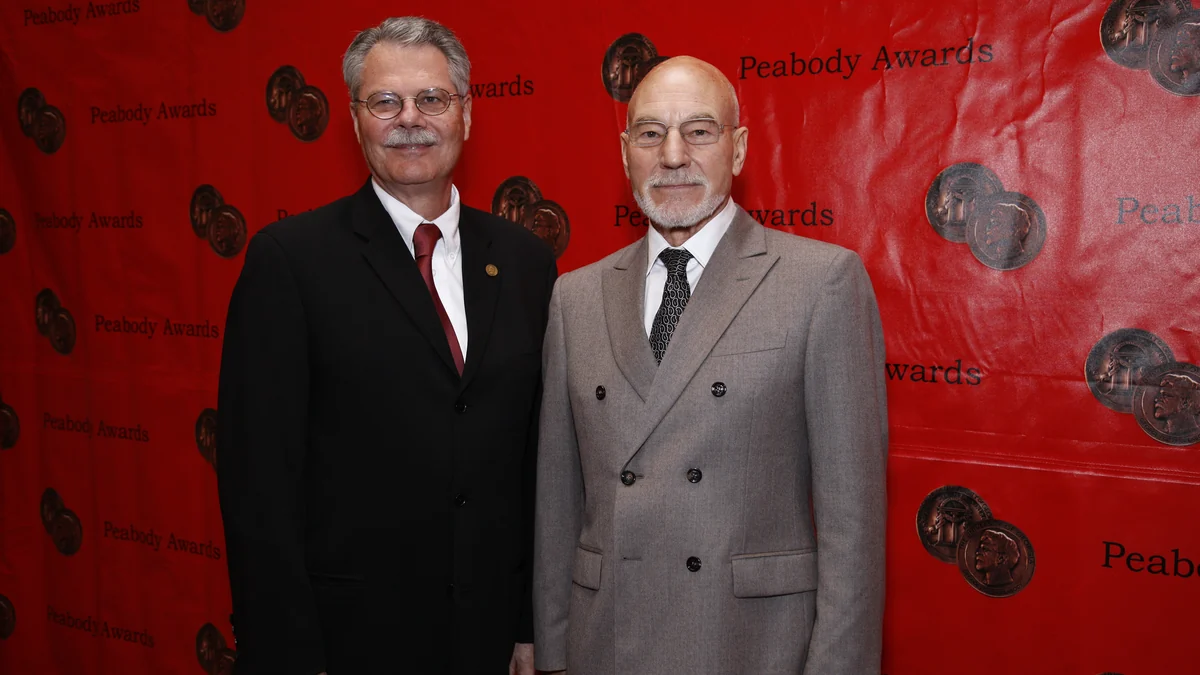 Patrick Stewart en costume gris aux côtés d’un autre homme devant le panneau rouge des Peabody Awards
