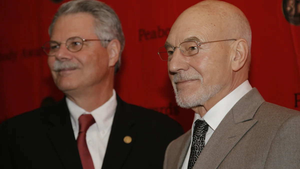 Patrick Stewart portant des lunettes et un costume gris au premier plan devant un fond rouge