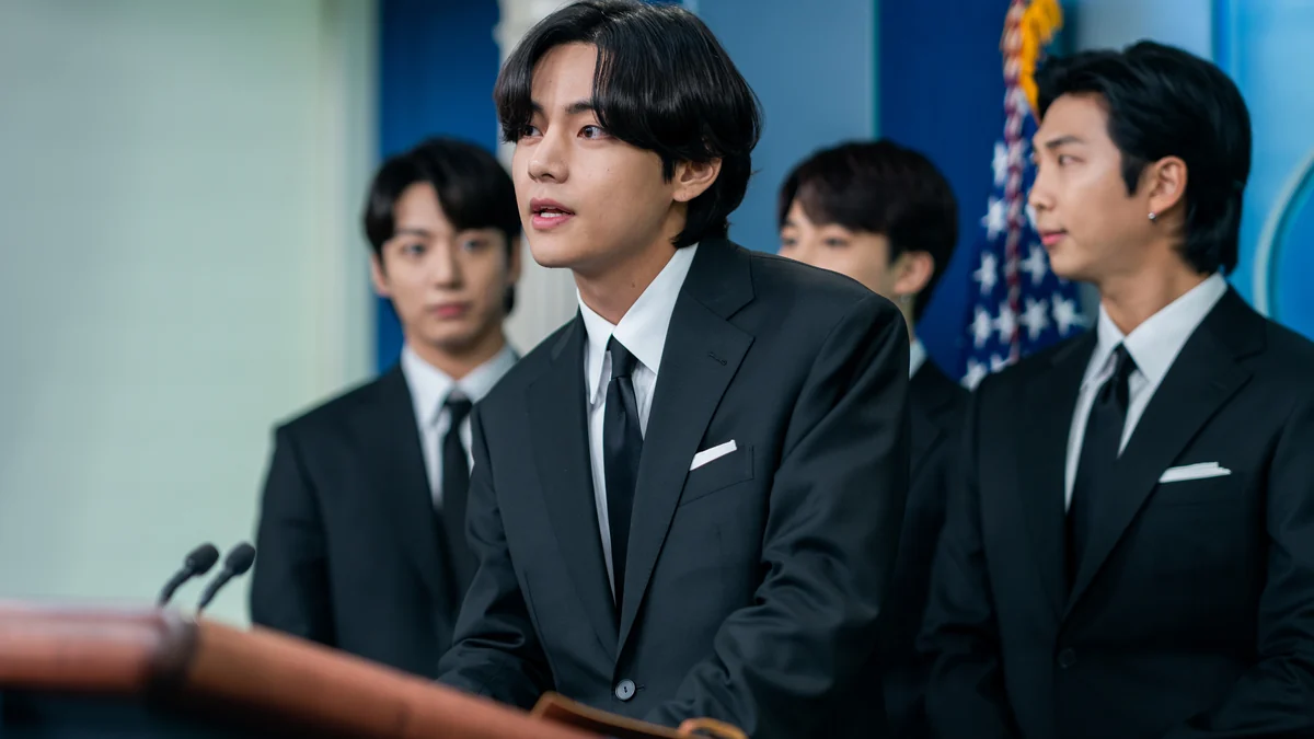 V, do BTS, fala diante de microfones, com outros integrantes do grupo ao fundo