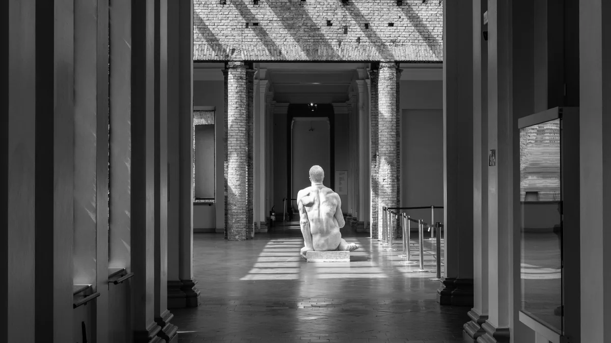 Galeria interna em preto e branco, com colunas de tijolos, feixes de luz e escultura de figura sentada ao centro