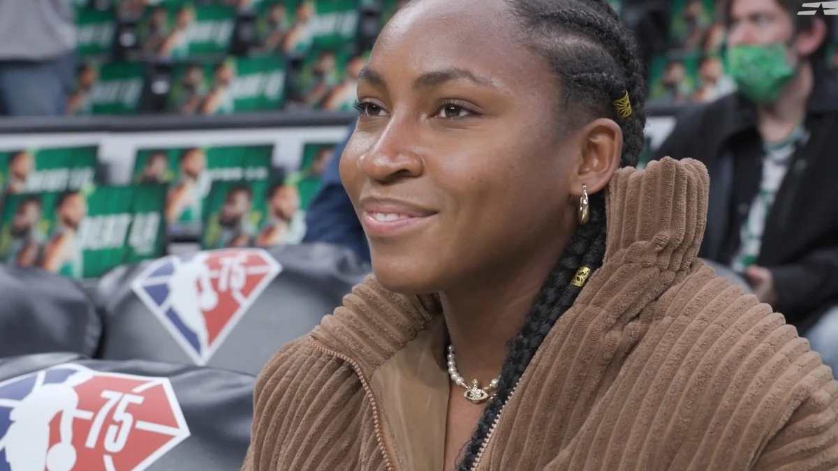 Coco Gauff seated in the stands, wearing a brown jacket and pearl necklace