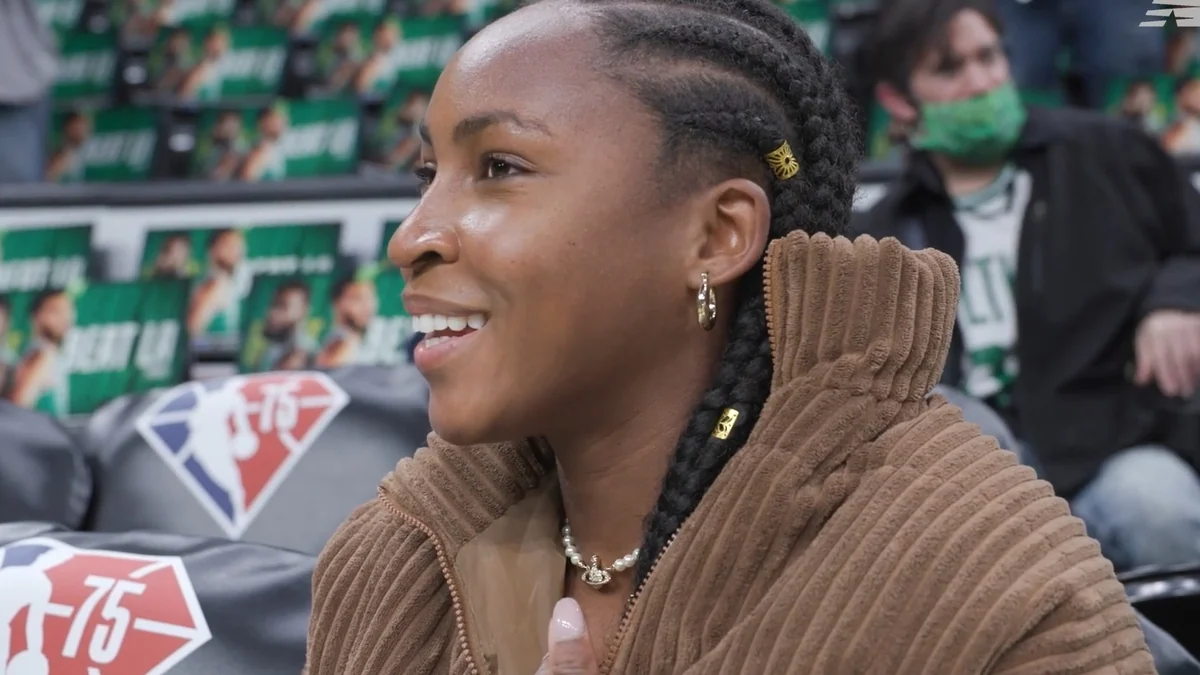 Coco Gauff smiling in the stands while wearing a brown jacket
