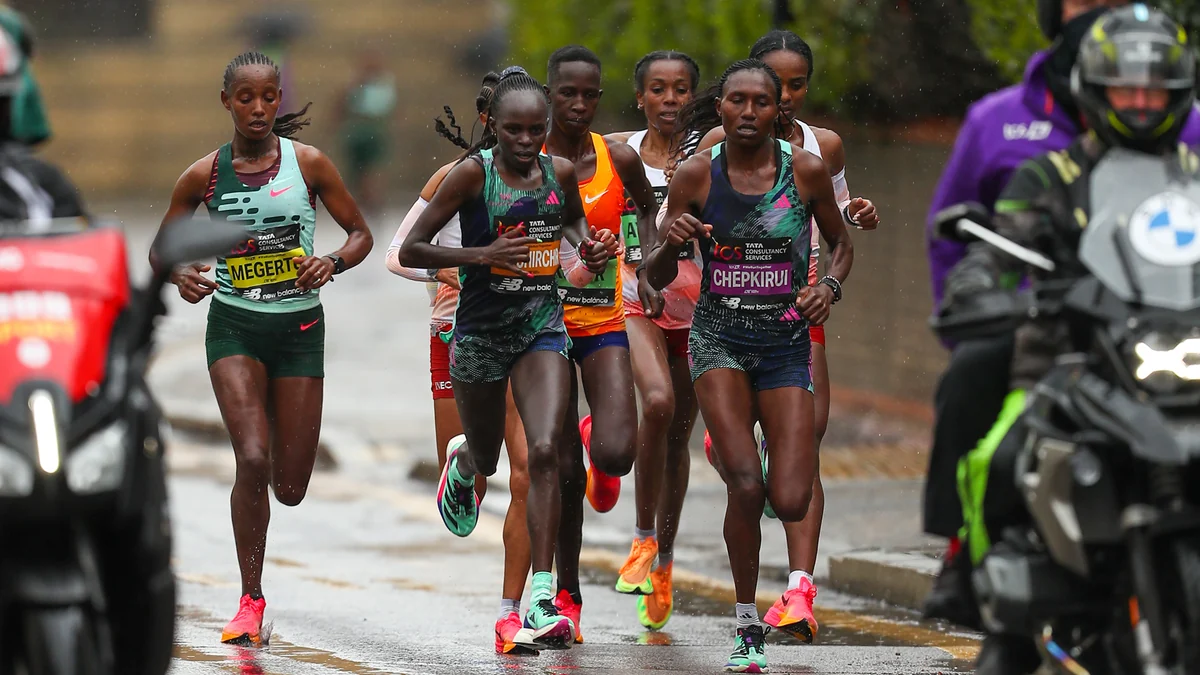 A group of elite women runners advances along a wet street, accompanied by motorcycles.