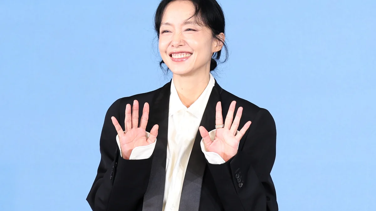 Jeon Do-yeon sorri e acena com as duas mãos diante de um fundo azul-claro.