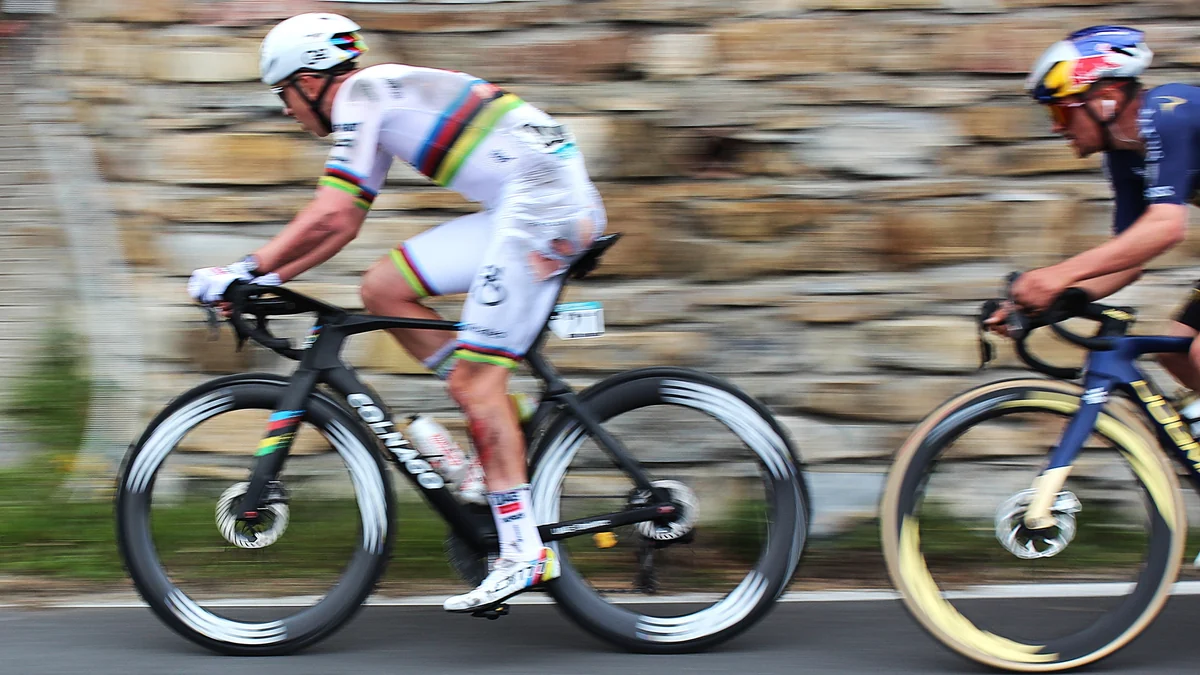 Tadej Pogačar in a white world champion’s kit rides ahead of another cyclist beside a stone wall.