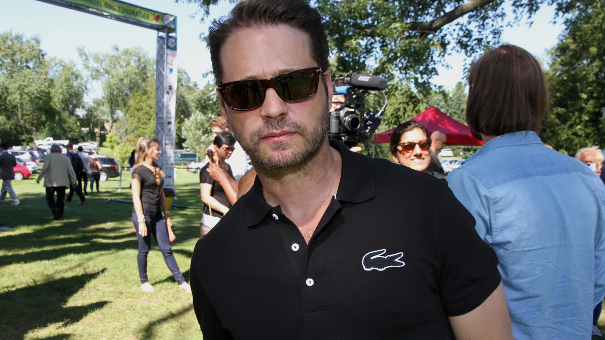 Jason Priestley porte des lunettes de soleil et un polo noir lors d’un événement en plein air