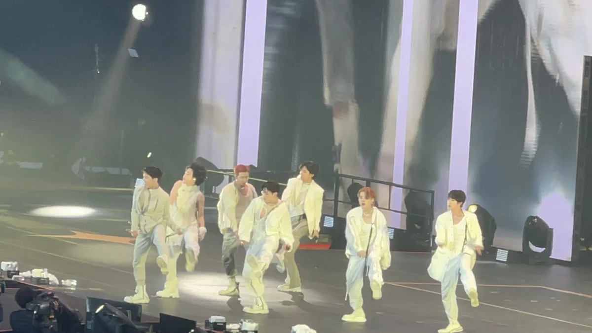 Sete integrantes do BTS se apresentam no palco com figurinos brancos diante de grandes painéis.